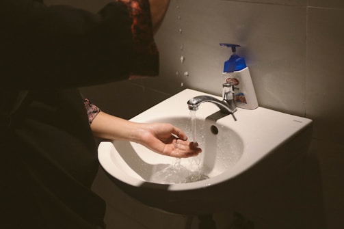 A person washing their hands under a running faucet in a sink. A liquid soap dispenser is positioned nearby on the sink's edge. Water is visibly splashing as it flows over the hands.
