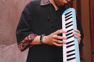 A person plays a melodica, wearing a black garment with red and gold patterned cuffs. The instrument has a white and black keyboard and a blue edge. The scene is set against a textured wall with muted tones.