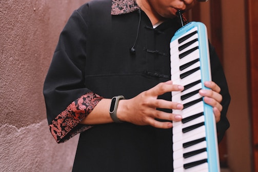 A person plays a melodica, wearing a black garment with red and gold patterned cuffs. The instrument has a white and black keyboard and a blue edge. The scene is set against a textured wall with muted tones.