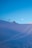A serene alpine landscape with a snow-covered slope under a clear blue sky. Ski lift chairs are visible on the left near a small wooden cabin, and ski tracks create gentle curves in the snow. The moon is seen in the sky above the scene.