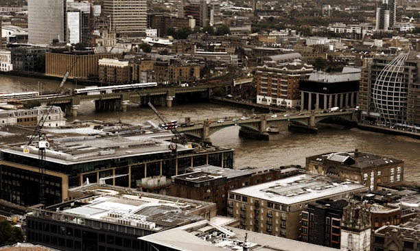 A bustling urban landscape features an array of modern and historical buildings clustered closely together. A river cuts through the city, with several bridges spanning across it, and there are visible cranes indicating ongoing construction. The scene captures a city's mix of architectural styles and signs of development.
