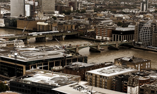 A bustling urban landscape features an array of modern and historical buildings clustered closely together. A river cuts through the city, with several bridges spanning across it, and there are visible cranes indicating ongoing construction. The scene captures a city's mix of architectural styles and signs of development.
