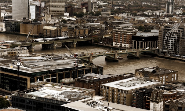A bustling urban landscape features an array of modern and historical buildings clustered closely together. A river cuts through the city, with several bridges spanning across it, and there are visible cranes indicating ongoing construction. The scene captures a city's mix of architectural styles and signs of development.