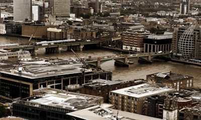 A bustling urban landscape features an array of modern and historical buildings clustered closely together. A river cuts through the city, with several bridges spanning across it, and there are visible cranes indicating ongoing construction. The scene captures a city's mix of architectural styles and signs of development.
