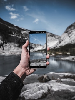 A hand holding a smartphone in front of a scenic landscape with mountains, sky, and a lake. The phone's screen shows the same landscape, creating a mirrored effect. The surroundings are rocky with clear water and the sky has scattered clouds.