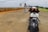 A motorcycle with a visible license plate is parked on a wide, empty road. A person wearing a helmet and a checkered shirt stands in front of the motorcycle, near a warning sign along a concrete barrier to the left. The background features a mix of cloudy skies and green hills.