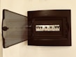 A black electrical panel is mounted on a wall with its transparent door open. Inside, there are several circuit breakers lined up in a row, each with visible labels and switches.