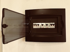A black electrical panel is mounted on a wall with its transparent door open. Inside, there are several circuit breakers lined up in a row, each with visible labels and switches.