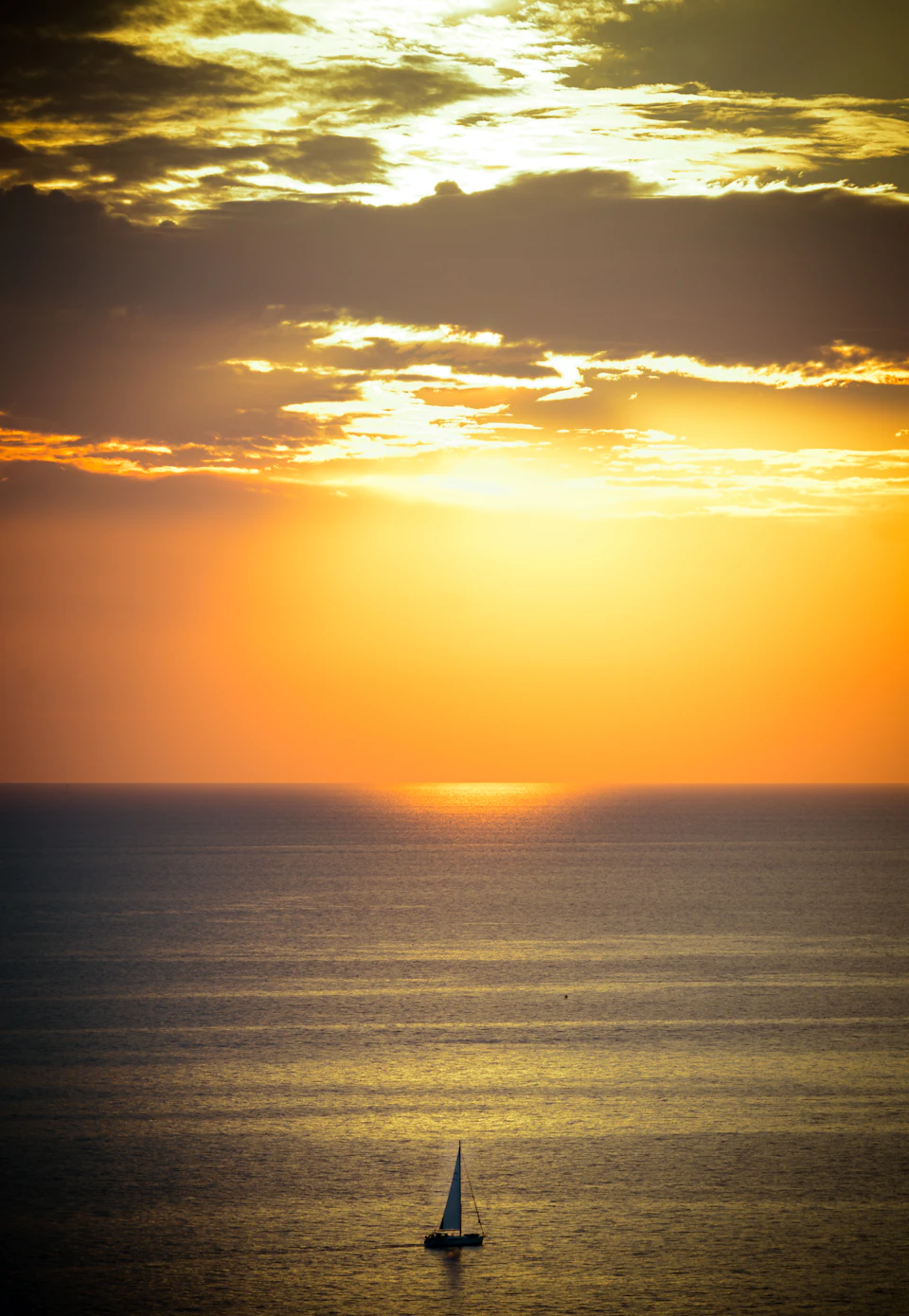 The image depicts a serene sunset over the ocean, with a sailboat gently floating on the water under a sky filled with clouds and hues of orange and yellow. The boat is positioned towards the right side of the frame, creating a sense of depth in the scene. The colors are vibrant, reflecting off the calm waters below.