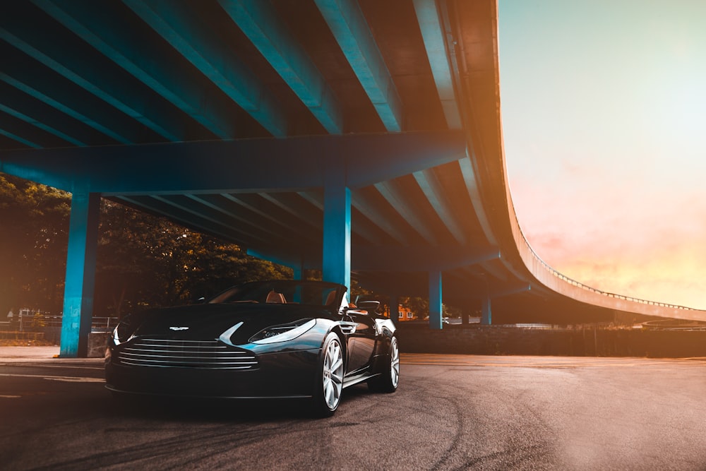 schwarzes Aston Martin Cabriolet Coupé parkt neben blauer Betonbrücke