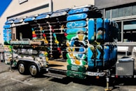 A creatively designed trailer structure made entirely from colorful skateboard decks. The trailer features vibrant blue, green, and multicolored surfaces, forming the sides and roof. It is parked outside a building with industrial-style windows, and there is a signboard attached to the trailer.
