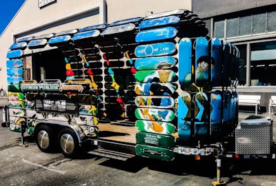A creatively designed trailer structure made entirely from colorful skateboard decks. The trailer features vibrant blue, green, and multicolored surfaces, forming the sides and roof. It is parked outside a building with industrial-style windows, and there is a signboard attached to the trailer.
