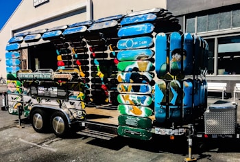 A creatively designed trailer structure made entirely from colorful skateboard decks. The trailer features vibrant blue, green, and multicolored surfaces, forming the sides and roof. It is parked outside a building with industrial-style windows, and there is a signboard attached to the trailer.