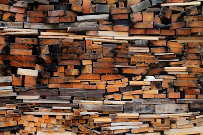 A large stack of wooden planks piled together in a somewhat disorganized manner. The wood varies in size, shape, and color, ranging from light to dark hues. Some pieces appear rough with visible grain patterns and others have smooth, sawed surfaces. The overall composition showcases a rustic and natural texture.