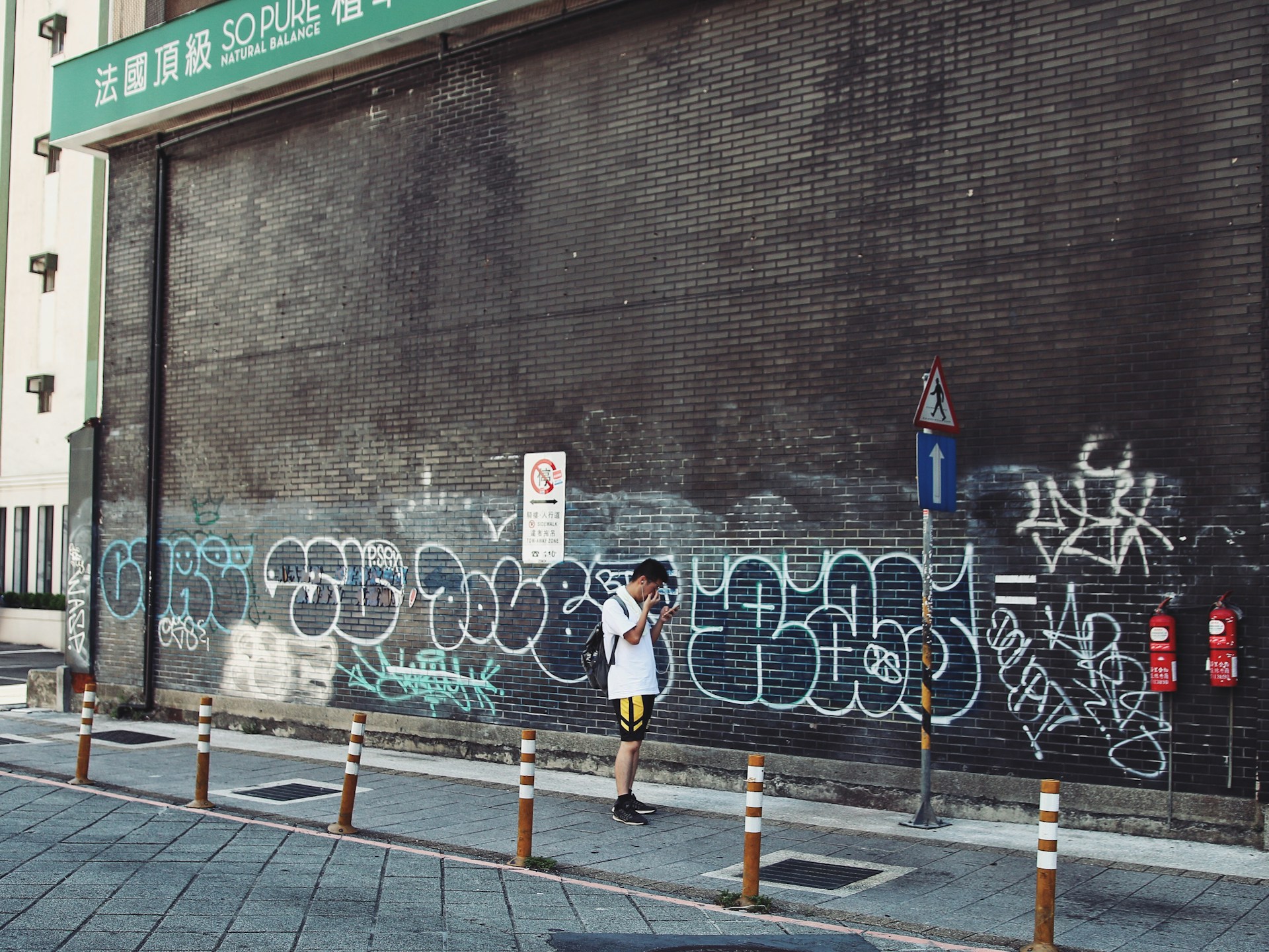 A person wearing casual sportswear stands on a street beside a building wall covered in various graffiti. The scene includes a few road signs and orange-striped bollards positioned along the sidewalk. The wall is large and dark, with white, blue, and green graffiti, while a green signboard with Chinese and English text is visible above.