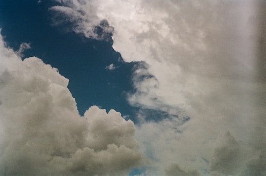 A sky filled with fluffy clouds, partially obscuring a blue background. The clouds exhibit varying shades of white and gray, indicating different densities and possibly different weather conditions.