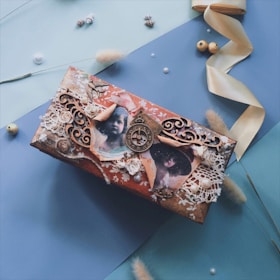 A rectangular vintage-style box on a two-tone blue background, adorned with ornate lace, sepia photographs of children, a decorative clock face, and floral embellishments. Surrounding the box are pastel-colored ribbons, seashells, small pearl-like beads, and dried grasses, adding to the nostalgic and artistic ambiance.