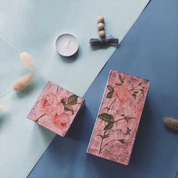 Two rectangular boxes with floral designs are placed on a surface divided into pastel green and blue areas. Nearby, a small white candle sits to the left of the boxes. On the right, a set of beige beads tied with a ribbon lies parallel to the candle. Fluffy beige plants or ornaments are scattered around, adding a textured element to the composition.