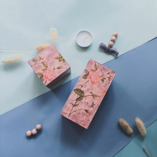 Two decorative boxes with floral patterns featuring roses are placed on blue and mint green surfaces. Alongside the boxes, there are small objects such as a tealight candle, fluffy white pampas grass stems, and wooden beads, creating a calm and artistic arrangement.