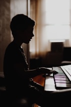 A silhouette of a child playing a keyboard in a dimly lit room. The light from the window casts a gentle glow on the curtains and the surroundings. The child's profile is visible, showing concentration and focus on the music.