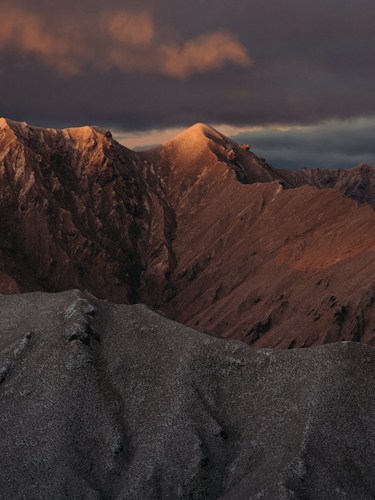 A dramatic mountain landscape with rugged peaks catching the warm light of the setting or rising sun. The sky is filled with dark, moody clouds, adding a sense of depth and atmosphere to the scene.
