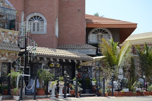 A detailed view of an ornate house with a mix of brick and stone architecture. The building features large arched windows, a spiral staircase, and a patio area adorned with numerous potted plants. A variety of decorative lamps can be seen near the entrance, and palm trees add a touch of greenery to the setting.