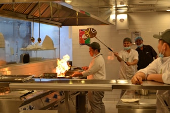 A busy commercial kitchen scene with chefs engaged in cooking. Flames rise from a pan held by one of the chefs as others look on and smile, indicating a lively and dynamic atmosphere. The kitchen features stainless steel surfaces, hanging utensils, and neatly arranged equipment.