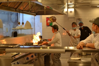 A busy commercial kitchen scene with chefs engaged in cooking. Flames rise from a pan held by one of the chefs as others look on and smile, indicating a lively and dynamic atmosphere. The kitchen features stainless steel surfaces, hanging utensils, and neatly arranged equipment.