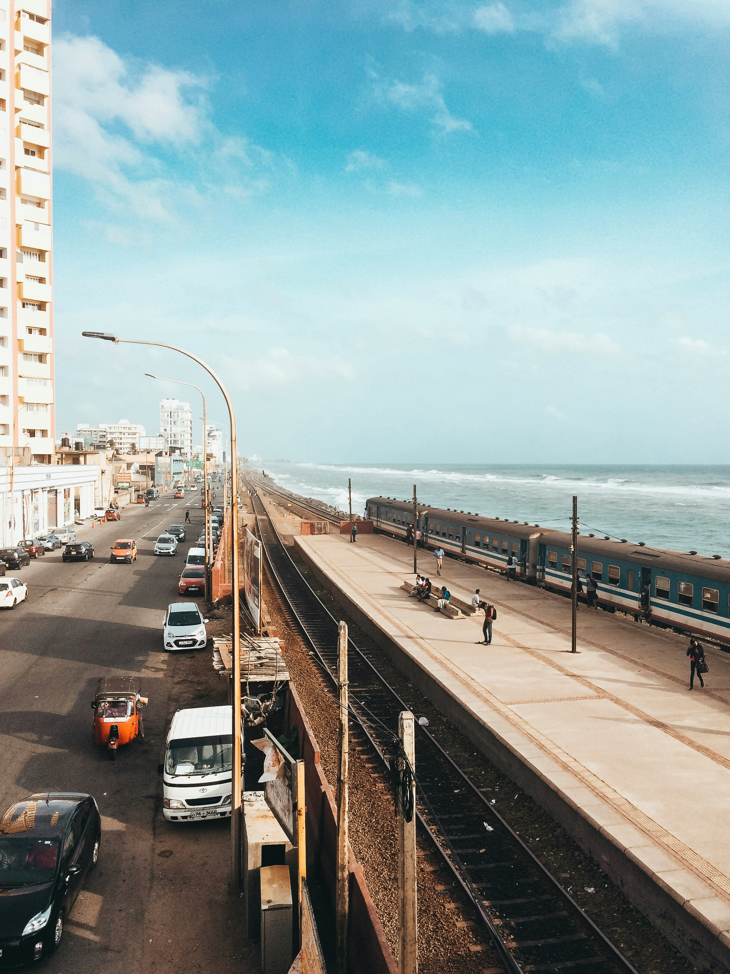 A photo of Marine Drive, Colombo. A train track lies between the sea and a road. Cars and a train drive by while people sit by the train tracks. The sky is light blue and the waves are calm.