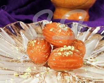 Three orange pastries garnished with crushed pistachios are placed on an intricately designed glass plate, surrounded by a purple cloth and a wooden item in the background.