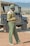 A man in a green uniform stands next to an open-top safari vehicle in a vast, open landscape. He appears to be engaged in an activity or conversation, with his hands together.