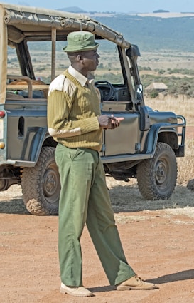 A man in a green uniform stands next to an open-top safari vehicle in a vast, open landscape. He appears to be engaged in an activity or conversation, with his hands together.