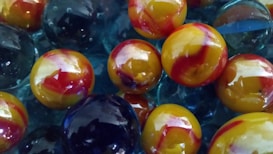 A close-up view of several glass marbles featuring vibrant colors. The marbles have a glossy finish, with swirling patterns of yellow and red on some, while others are a deep blue color. They are clustered together, creating a dynamic composition with reflective light highlights.