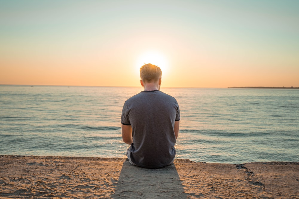 man sitting on sand front of sea during golden hour