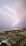 A dramatic scene of lightning striking over a body of water in the distance. The foreground features an urban park with a large, modern, open-air pavilion and surrounding greenery. Numerous small lights outline paths and trees, adding a contrast to the dark landscape.