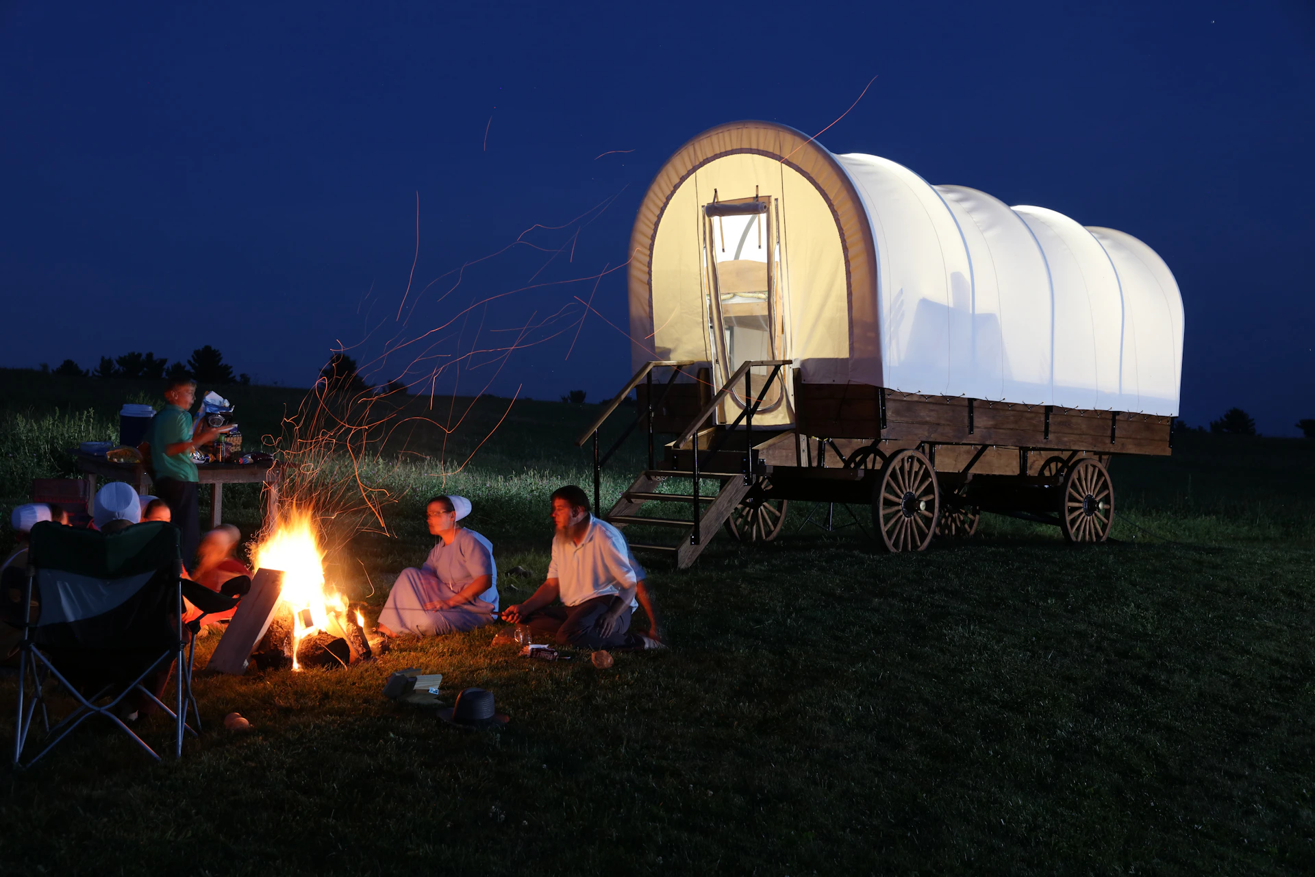 A group of riders gathered around a campfire at dusk on a rustic ranch.