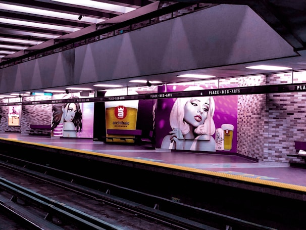 A subway platform with vibrant wall murals depicting stylized portraits of women with distinctive makeup and hair. The murals are complemented by an advertisement for a microbrewery, featuring a glass of beer. The platform is well-lit and lined with benches beneath a grid of overhead lights, with metro tracks visible in the foreground.