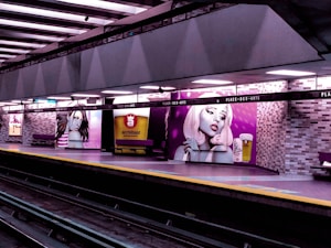 A subway platform with vibrant wall murals depicting stylized portraits of women with distinctive makeup and hair. The murals are complemented by an advertisement for a microbrewery, featuring a glass of beer. The platform is well-lit and lined with benches beneath a grid of overhead lights, with metro tracks visible in the foreground.