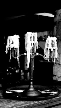 A vintage metal candelabrum with three holders, each adorned with melted and dripped wax, creating a dramatic and artistic effect. The background is dimly lit, emphasizing the intricate details of the wax and the aged texture of the candelabrum.