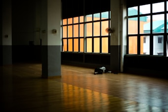 A large, empty indoor space with a wooden floor and large windows letting in natural light. There is a small pile of belongings or possibly a person resting on the floor near the windows. The room feels spacious with high ceilings and there is a noticeable contrast between the dim interior and the colorful buildings outside.