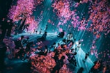 A visually immersive art installation features a group of people interacting with digital projections that create the effect of falling cherry blossoms. The space is filled with bright pink and purple petals against a backdrop of cascading blue light, providing a dreamlike atmosphere.