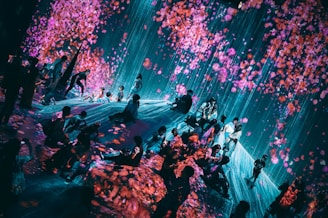 A visually immersive art installation features a group of people interacting with digital projections that create the effect of falling cherry blossoms. The space is filled with bright pink and purple petals against a backdrop of cascading blue light, providing a dreamlike atmosphere.