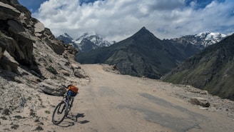 A bicycle with an orange bag is parked on a rugged mountain path. The surroundings feature rocky terrain with majestic mountains in the background, some of which are capped with snow. The sky is partly cloudy.