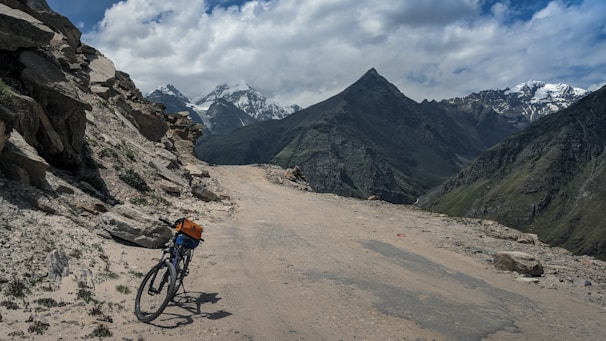 A bicycle with an orange bag is parked on a rugged mountain path. The surroundings feature rocky terrain with majestic mountains in the background, some of which are capped with snow. The sky is partly cloudy.