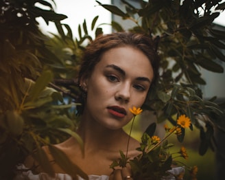 A woman with a serene expression is surrounded by lush green foliage. Her hair is brown and loosely tied back, and she wears a subtle, natural makeup look. She gently holds a small bouquet of vibrant yellow flowers in front of her chest.