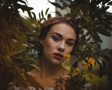 A woman with a serene expression is surrounded by lush green foliage. Her hair is brown and loosely tied back, and she wears a subtle, natural makeup look. She gently holds a small bouquet of vibrant yellow flowers in front of her chest.