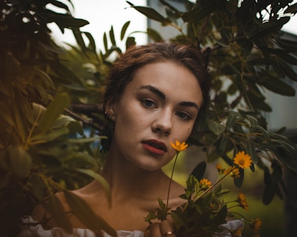 A woman with a serene expression is surrounded by lush green foliage. Her hair is brown and loosely tied back, and she wears a subtle, natural makeup look. She gently holds a small bouquet of vibrant yellow flowers in front of her chest.