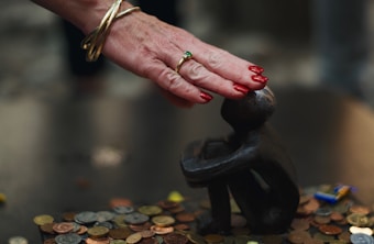 A hand with red painted nails and gold bangles gently touches the top of a small black statue of a crouching figure. The hand also has a green ring on the ring finger. The statue is placed on a surface covered with various coins.