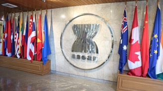 A row of diverse national flags is displayed, representing various countries, alongside a large ASEAN emblem mounted on a marble wall. The area is well-lit and designed with a polished wooden floor and ceiling.