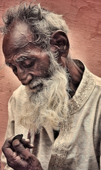 An elderly man with a long white beard gazes downwards pensively while holding a small dark object in his hand. His expression suggests contemplation or deep thought. He wears a textured garment adorned with intricate embroidery against a backdrop of soft, muted brown tones.